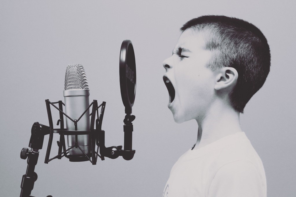 Image en noir et blanc montrant au premier plan un enfant qui semble crier dans un micro d'enregistrement (tel que ceux qu'on utilise pour enregistrer des morceaux de musique ou des livres audio dans des studios). L'enfant est de profil sur la droite de la photo ; le micro à gauche.