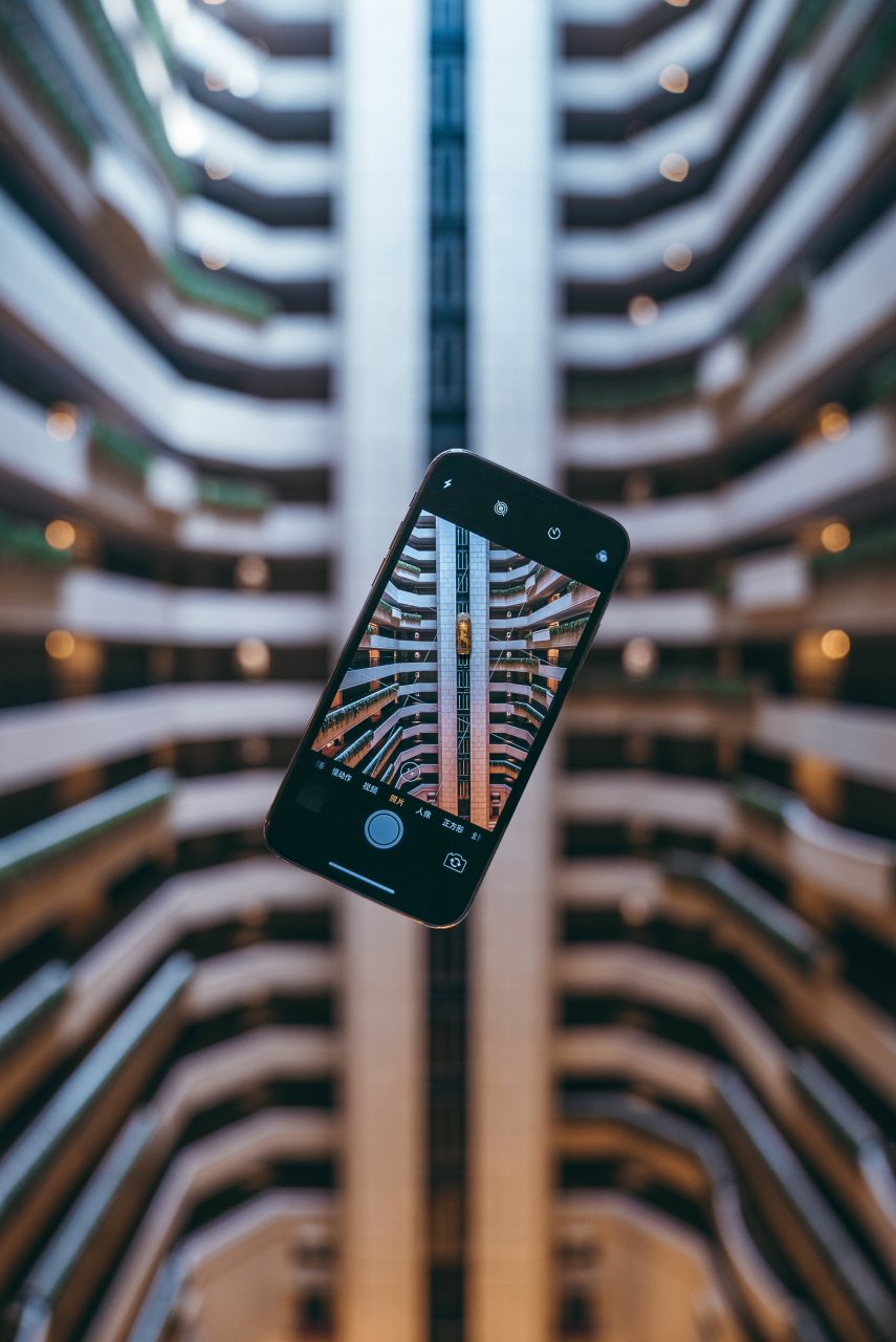 Photographie illustrant le principe d'une exposition virtuelle. Sur le fonds, une pièce d'architecture : un escalier ou des étages. Au premier plan, un smartphone en mode "photographie".