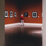 Photographie prise dans un musée. On y voit une personne seule au milieu d'une salle d'exposition aux murs rouges. Des oeuvres entourées de cadres blancs sont exposées.
