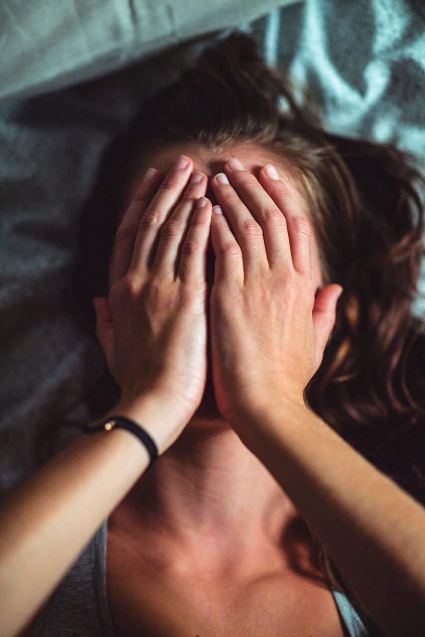 Photographie en couleur. Zoom sur le cou et le visage d'une jeune femme allongée sur un lit dont les draps sont froissés et bleus. Elle mets ses mains en coupole sur sa tête ; ce qui nous empêche de voir ses oreilles, sa bouche, ses yeux et son front.