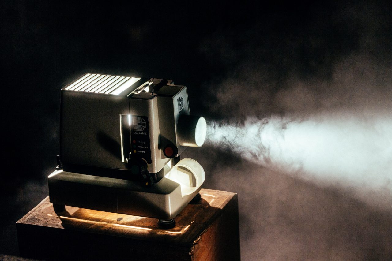 Photographie d'un projecteur en fonctionnement, pour illustrer le top 10 des films à visionner pendant le confinement. La machine, grise, est posée sur un tabouret en bois dans une pièce plongée dans l'obscurité. Un faisceau blanc sort du projecteur, projetant les images (que l'on ne voit pas dans le cadre de la photographie).