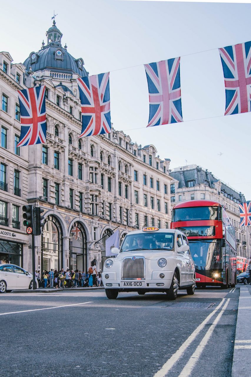 Photographie d'une rue dans la ville de Londres. Un taxi anglais et un bus à impériale roulent sous des bannières adorant le drapeau du Royaume-Uni.