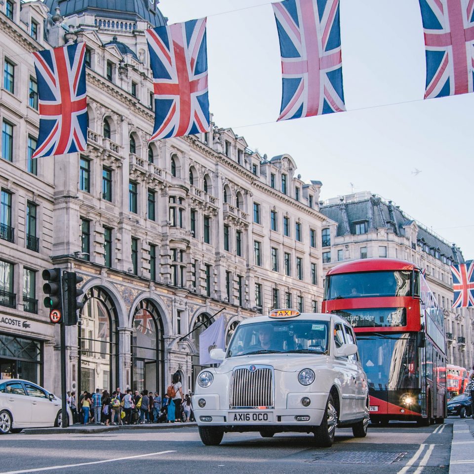 Photographie d'une rue dans la ville de Londres. Un taxi anglais et un bus à impériale roulent sous des bannières adorant le drapeau du Royaume-Uni.