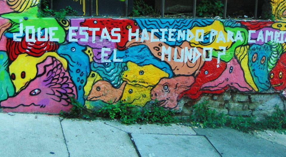 Photographie illustrant le thème "cultural industries". Mur coloré, peint avec une oeuvre de street art. Une écriture transparaît en blanc: que faites-vous pour changer le monde (sur la photo, en espagnol) ?